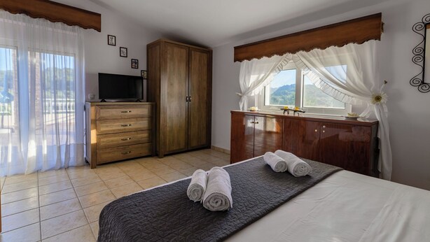 A bedroom with a bed, a wooden wardrobe, a television, and a window with curtains. - Laylande House with Private Pool (Theologos)