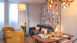 A modern living room with a dining area, featuring a sofa, chairs, a table set with breakfast, and a wall with abstract art. - Montmartre Paris Suite (Paris)