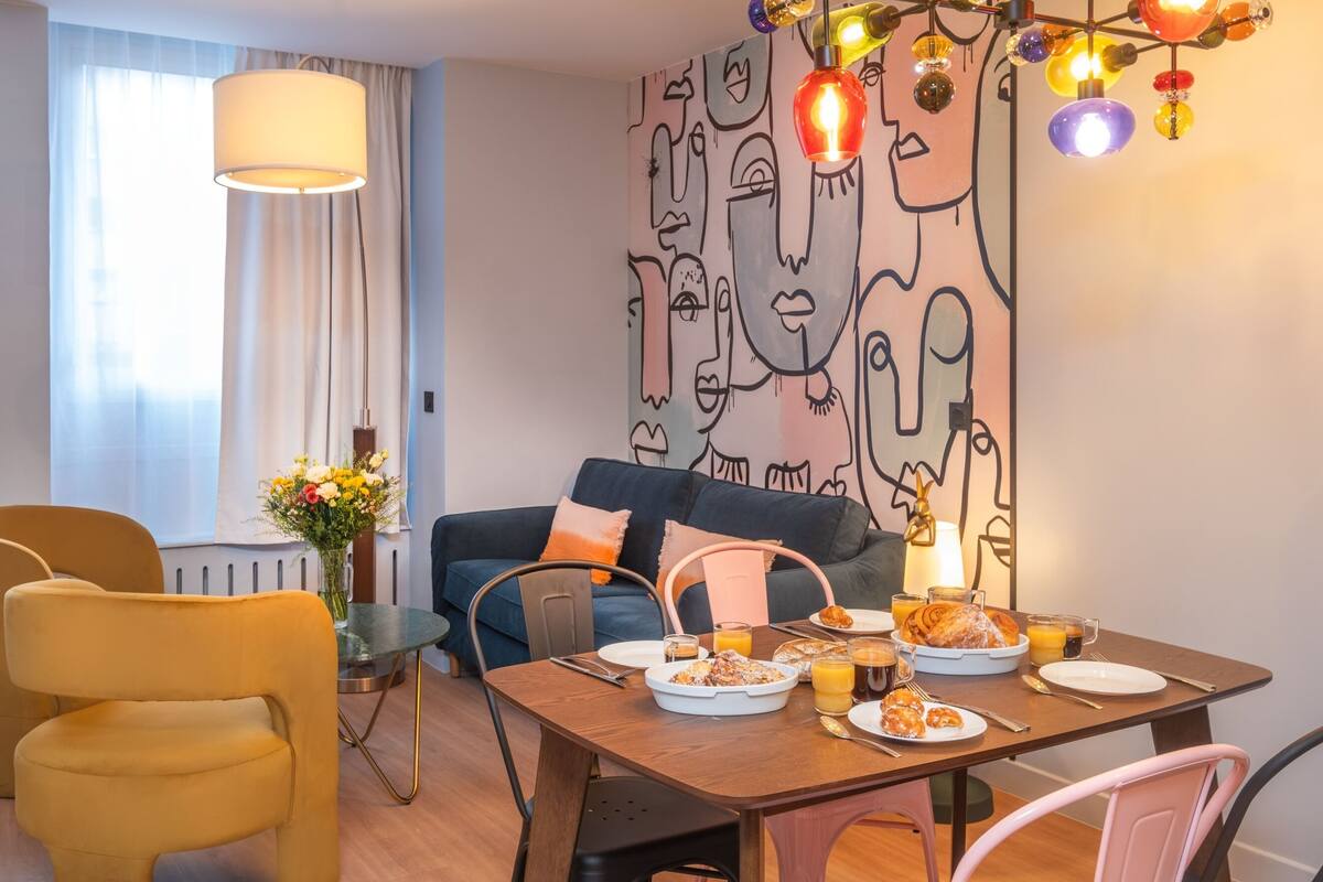 A modern living room with a dining area, featuring a sofa, chairs, a table set with breakfast, and a wall with abstract art.