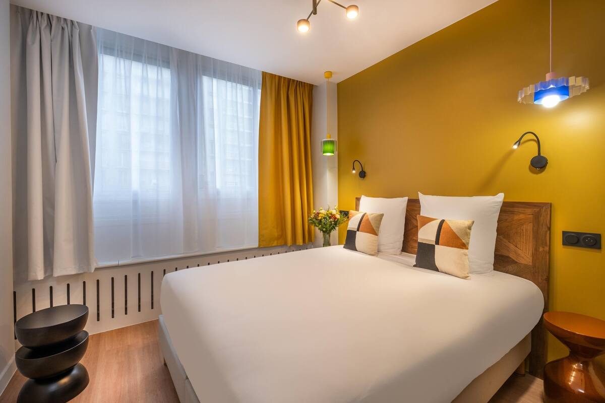A neatly made bed with white bedding and decorative pillows, a bedside table with a vase of flowers, and a modern pendant light.