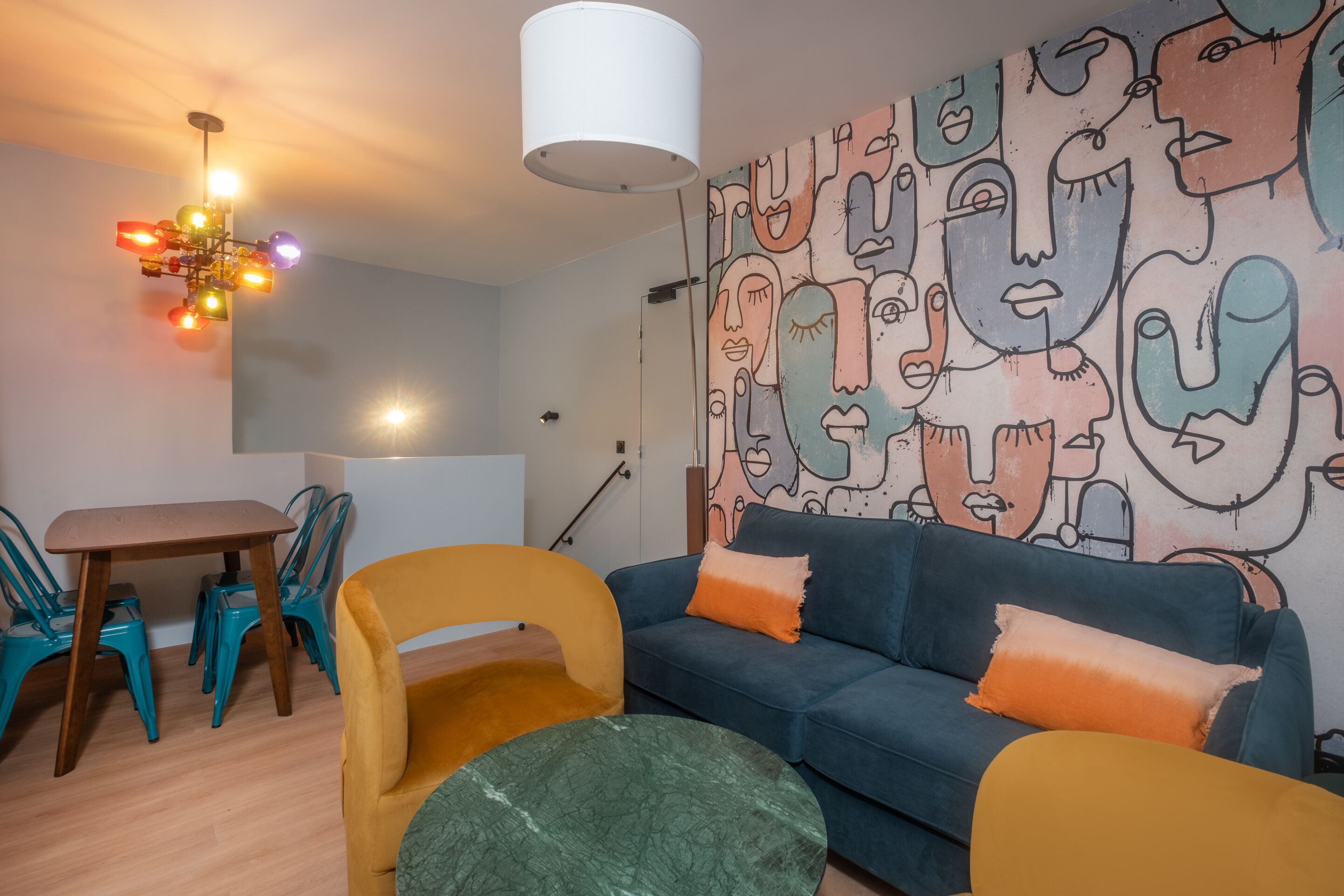 A modern living room with a blue sofa, yellow armchair, and teal chairs. A ceiling light fixture and a wall mural are also visible.