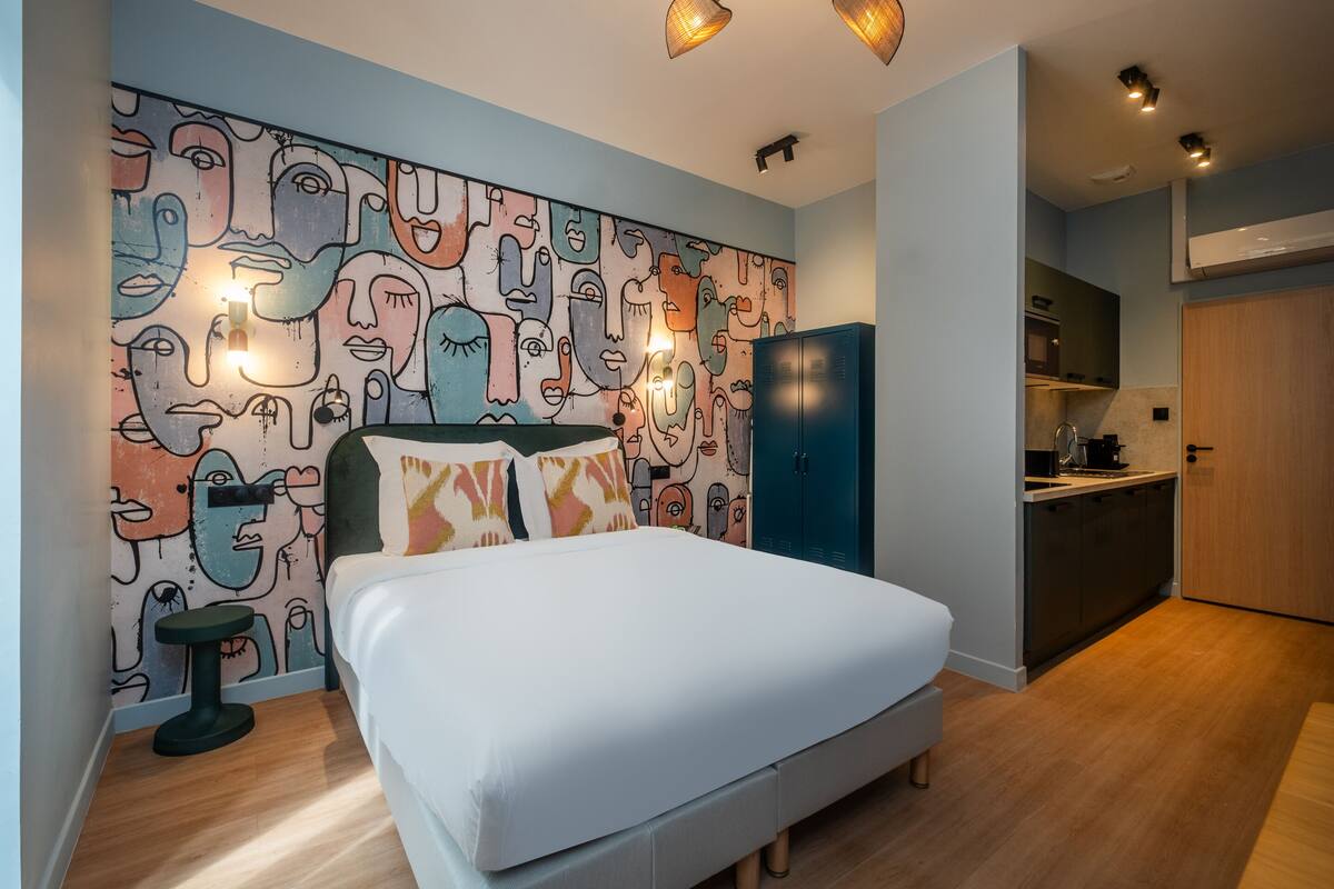 A bedroom with a bed, a nightstand, a wall with abstract art, a wardrobe, and a kitchenette in the adjacent room.