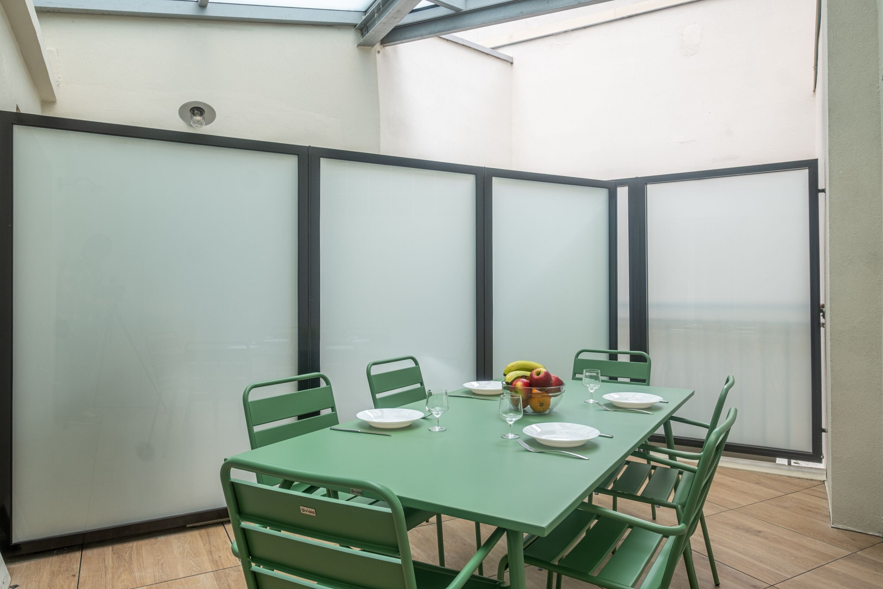 A dining area with a green table and chairs, a bowl of fruit, and a window with a view.