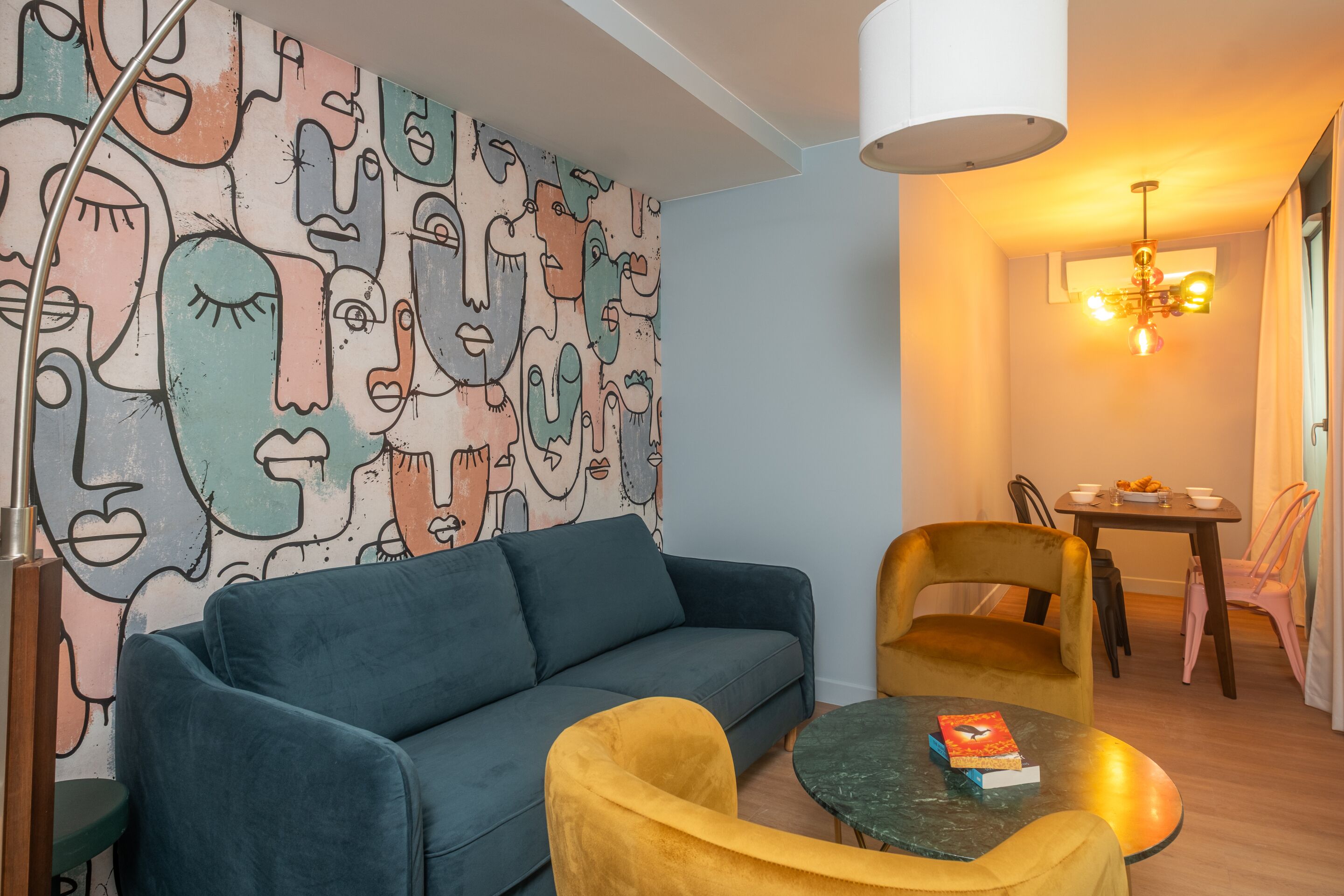 Una sala de estar moderna con un sofá azul, un sillón amarillo y una pequeña mesa redonda con un libro. Un mural abstracto y colorido adorna la pared, y un candelabro cuelga del techo.