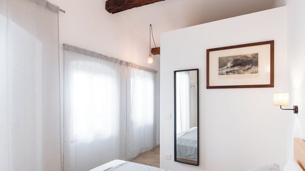 Une chambre à coucher comprenant un lit, une table de chevet, un miroir, un tableau encadré et une fenêtre avec des rideaux légers.