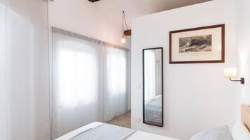 Una camera da letto con un letto, un comodino, uno specchio, un quadro incorniciato e una finestra con tende leggere.