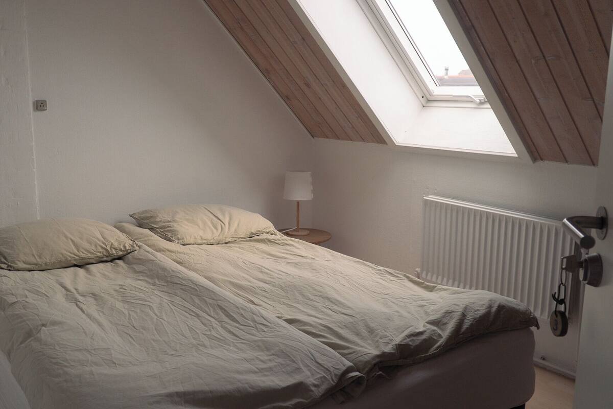 A bedroom with a sloped ceiling, a bed with beige bedding, a bedside lamp, and a radiator.