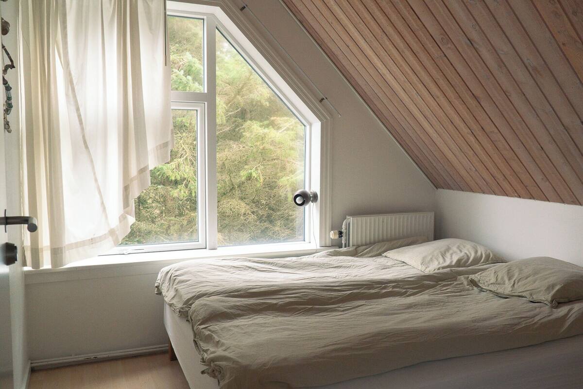 A bedroom with a sloped ceiling, a window with curtains, a radiator, and a bed with a beige blanket.