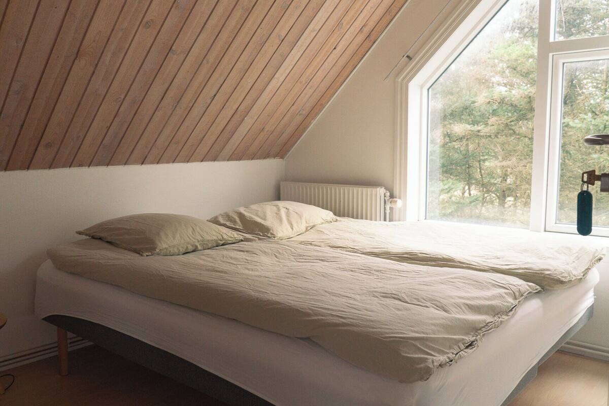 A bedroom with a sloped ceiling, a bed with beige bedding, a small wooden stool, a floor lamp, and a window with a view of trees.