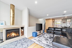 A modern living room with a fireplace, a flat-screen TV, a black leather sofa, and a staircase leading to an upper level.