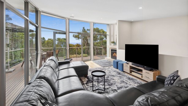 A modern living room with a large TV, leather sofa, and a balcony with a view. - Beachfront 63 (Lorne)