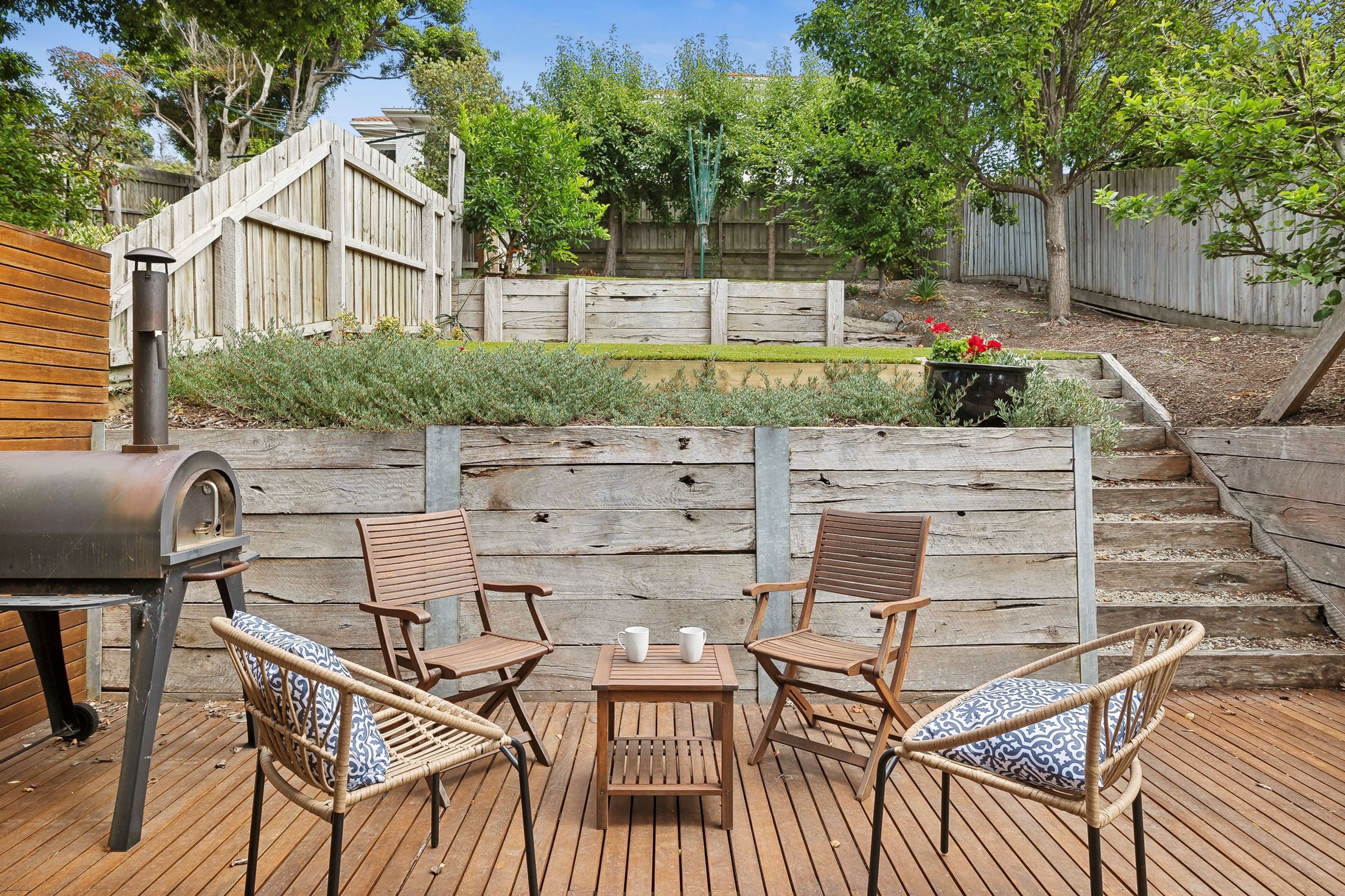 Une terrasse en bois avec deux chaises en osier et une petite table, un barbecue et une clôture en bois en arrière-plan.