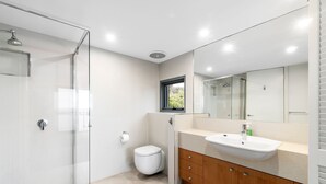 A modern bathroom with a glass shower enclosure, a toilet, a sink with a mirror above it, and a window providing natural light. - Beachfront 63 (Lorne)