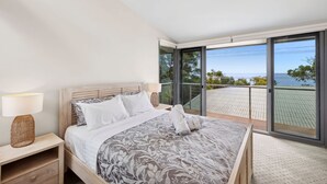 A bedroom with a large bed, a nightstand, a lamp, and a view of the sea through a sliding glass door. - Beachfront 63 (Lorne)