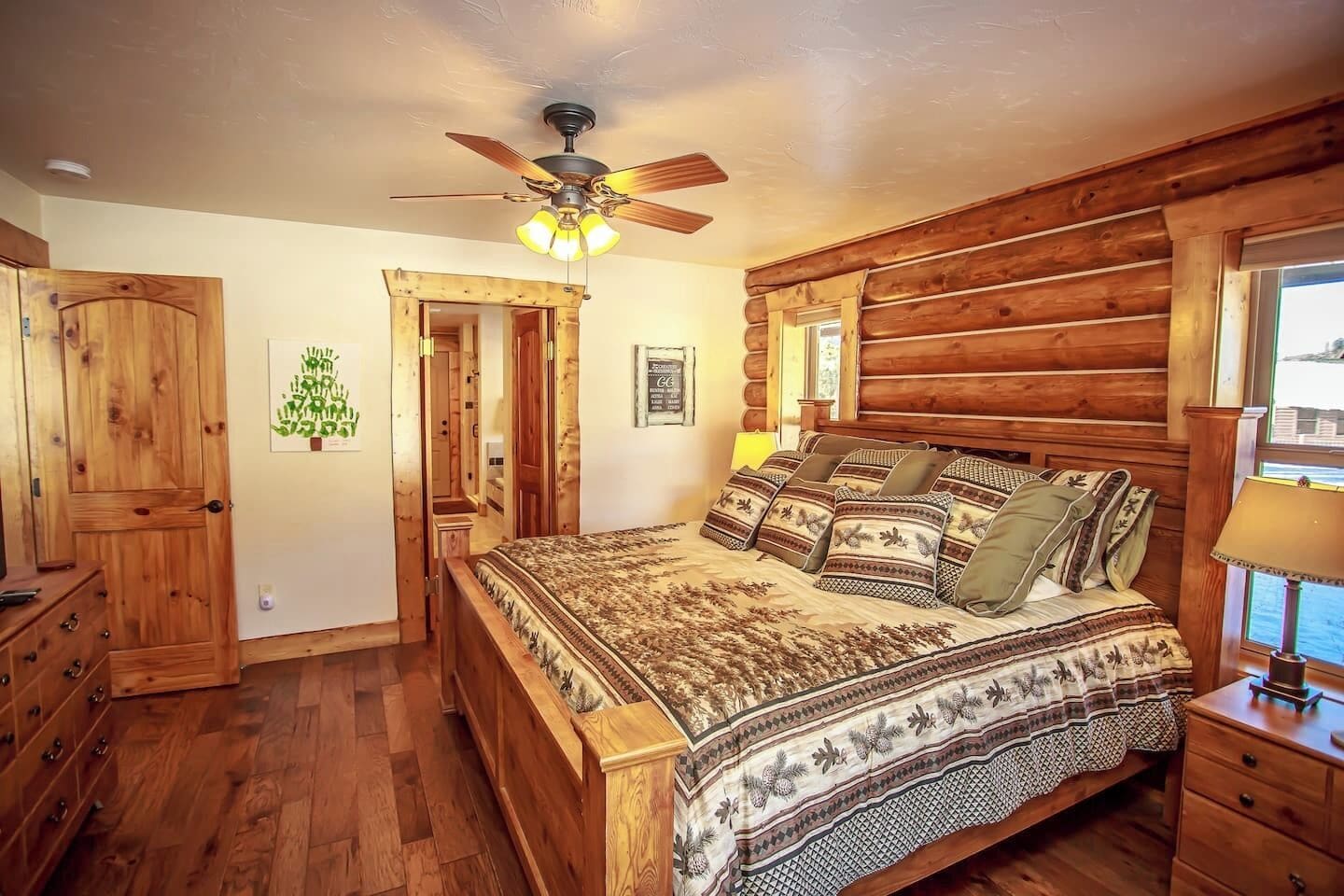 A log cabin-style bedroom with a bed, wooden walls, a ceiling fan, and a view through a window.