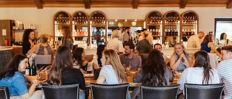 A restaurant with wooden beams on the ceiling, tables with chairs, and a bar area with shelves of bottles.