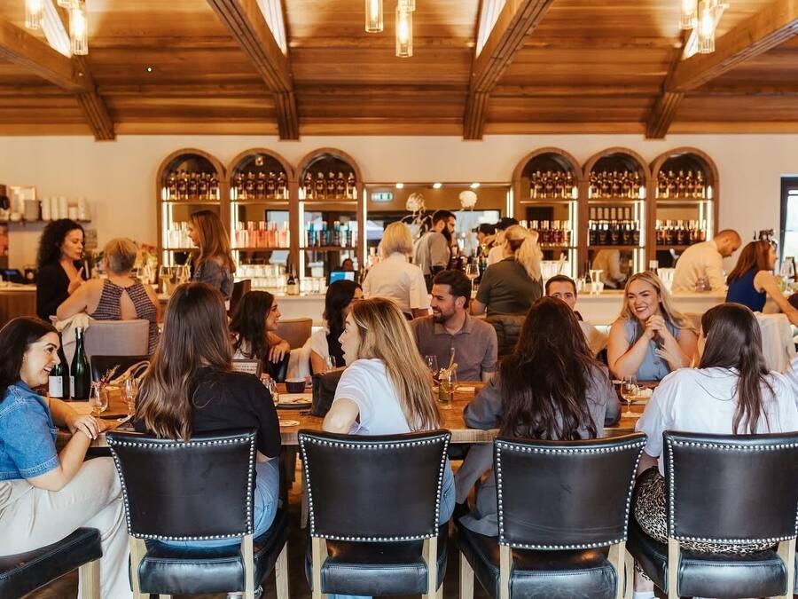 A restaurant with wooden beams on the ceiling, tables with chairs, and a bar area with shelves of bottles.