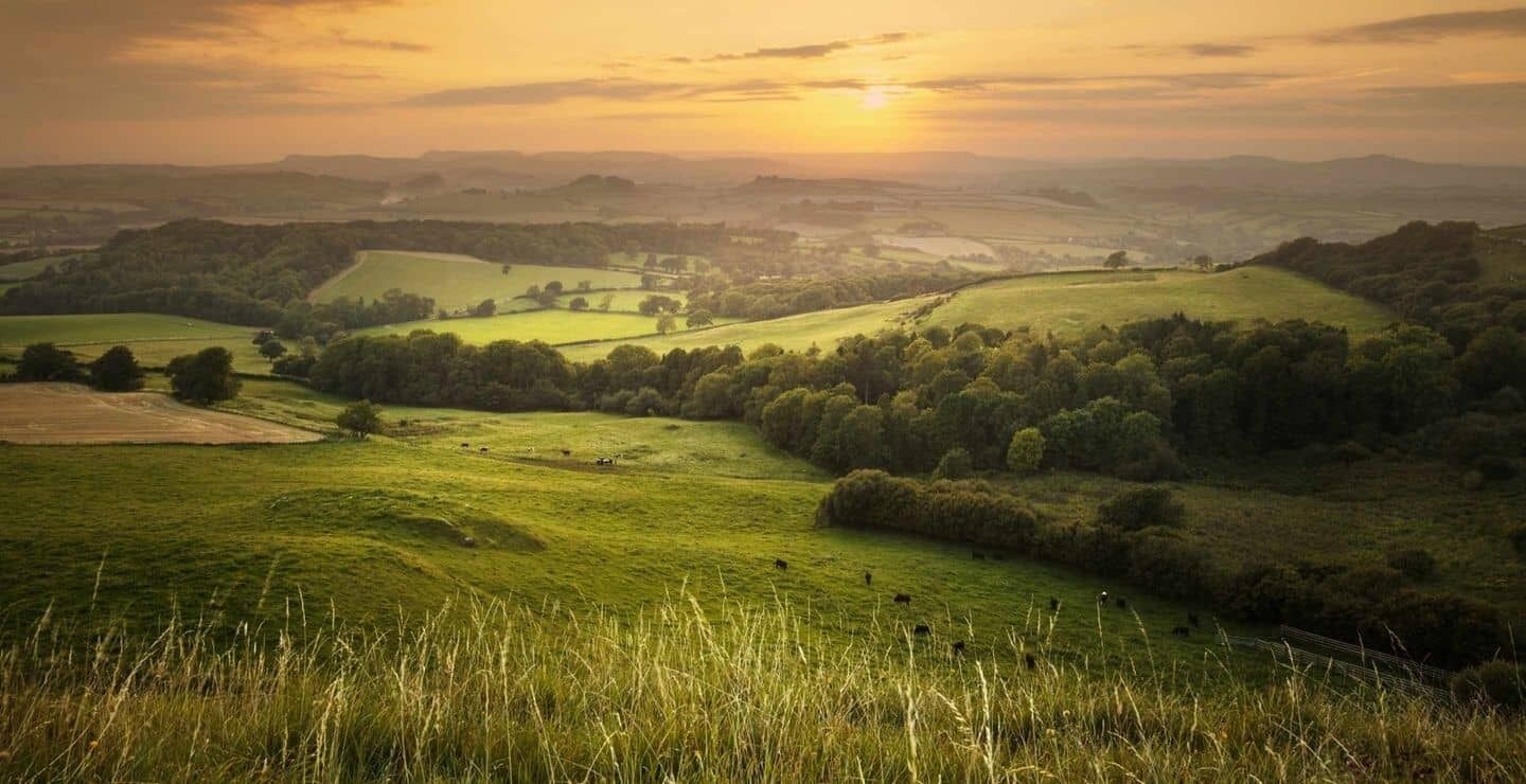 Paesaggio rurale con dolci colline, campi e un tramonto all'orizzonte.
