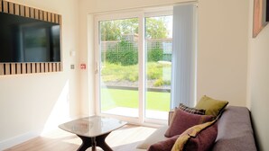 Una sala de estar moderna con una mesa de vidrio, un sofá con cojines, un televisor de pantalla plana montado en la pared y un ventanal corredizo que da a un jardín.