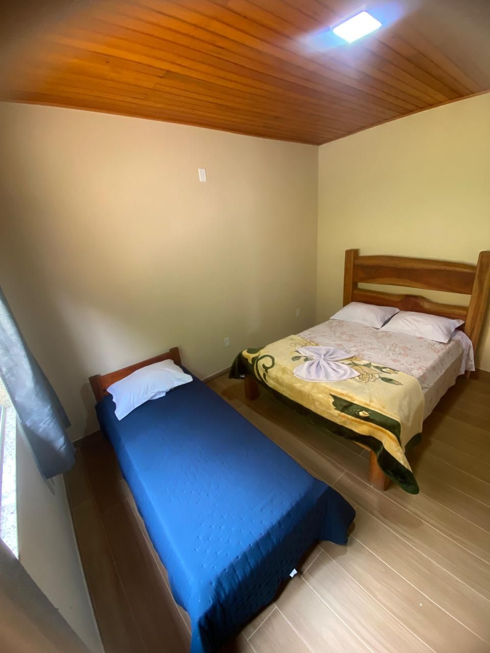 Ein Zimmer mit zwei Betten, eines mit blauer Bettdecke, das andere mit Blumenmuster. Der Raum hat Holzboden und eine Holzwand mit Oberlicht.