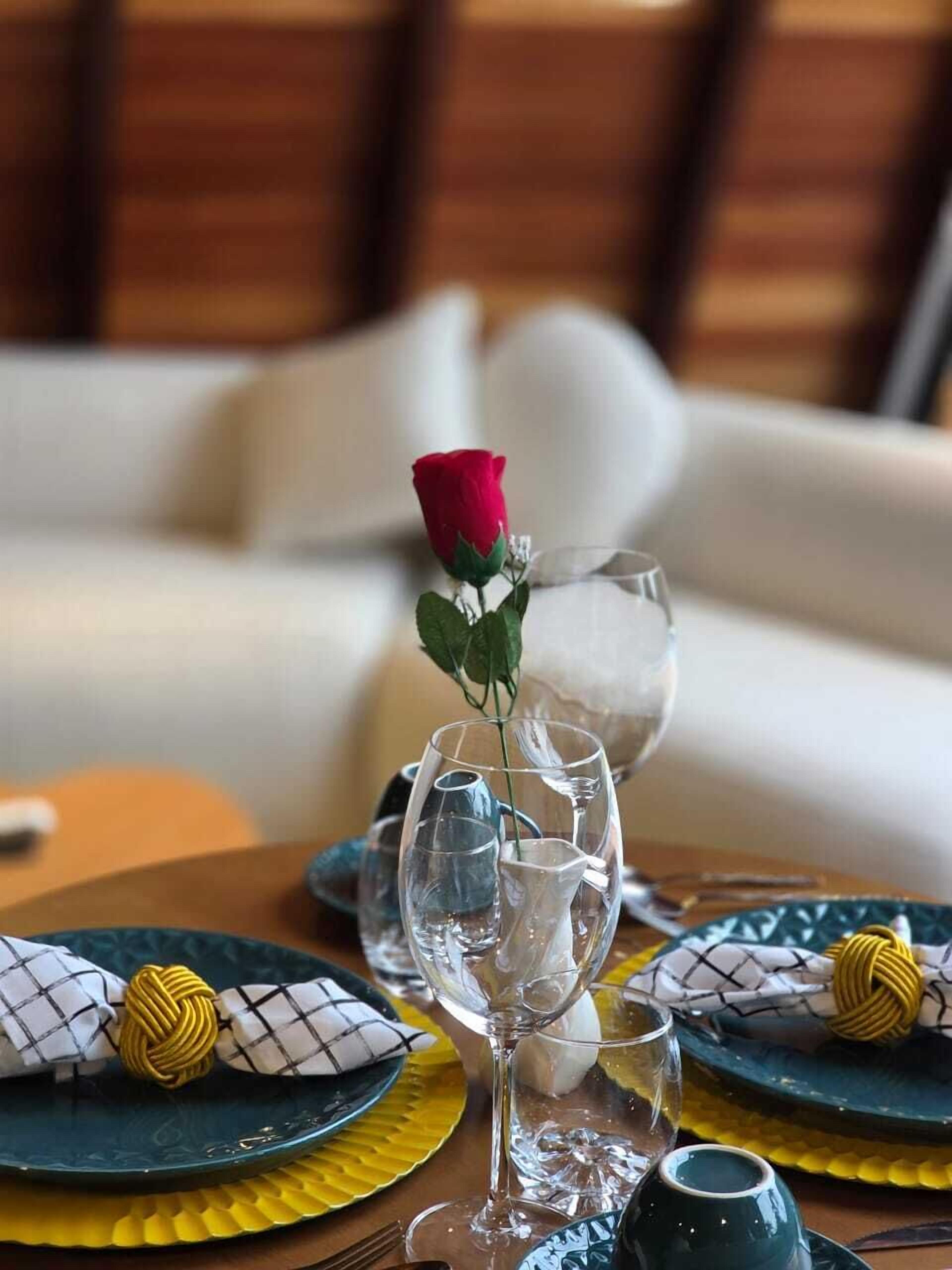 Uma mesa de jantar posta com copos de vidro transparente, uma única rosa vermelha e guardanapos estampados.