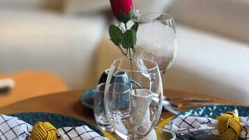 Uma mesa de jantar posta com copos de vidro transparente, uma única rosa vermelha e guardanapos estampados.