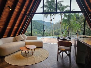 A modern interior with a large window overlooking a lush landscape, a sofa, a dining table, and a bar area. - Delícias da Tilápia (Domingos Martins)
