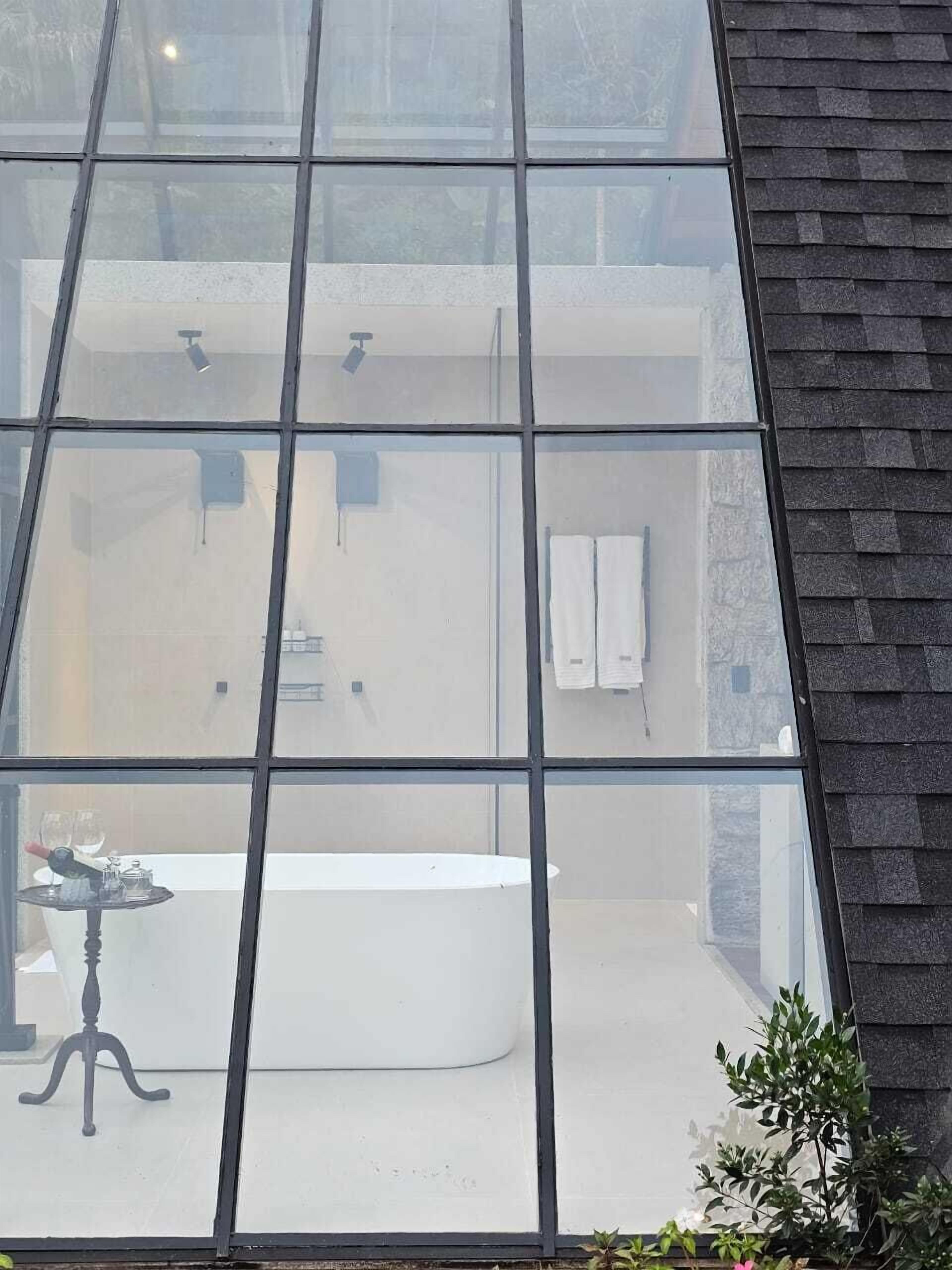 Un baño moderno con una bañera grande, un lavamanos y un inodoro. Hay una ventana con diseño de cuadrícula, y una mesita con una botella y un vaso.