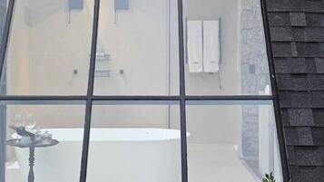 Un baño moderno con una bañera grande, un lavamanos y un inodoro. Hay una ventana con diseño de cuadrícula, y una mesita con una botella y un vaso.