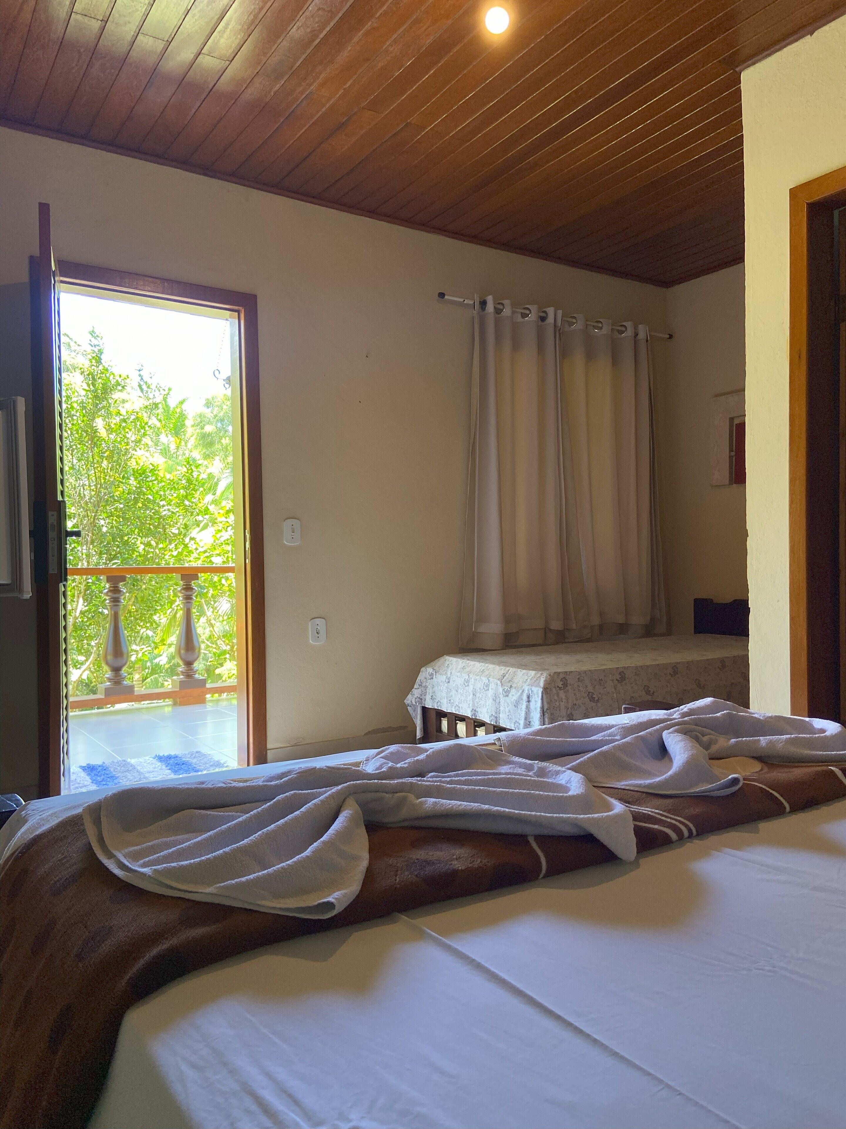 A bedroom with a bed, a window with curtains, a view of greenery outside, and a wooden ceiling.