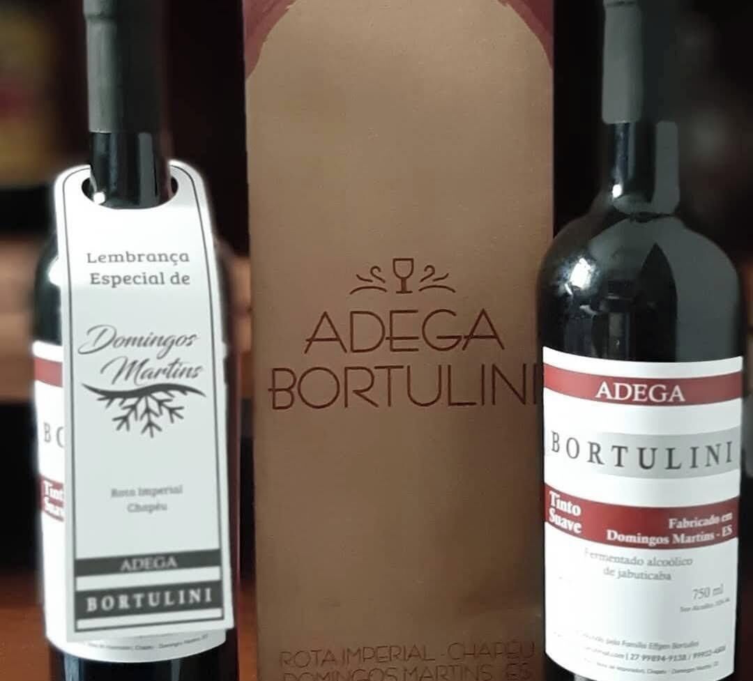 Three bottles of Adegá Bortulini wine, one with a white label and two with red labels, placed in front of a brown box with the Adegá Bortulini logo.