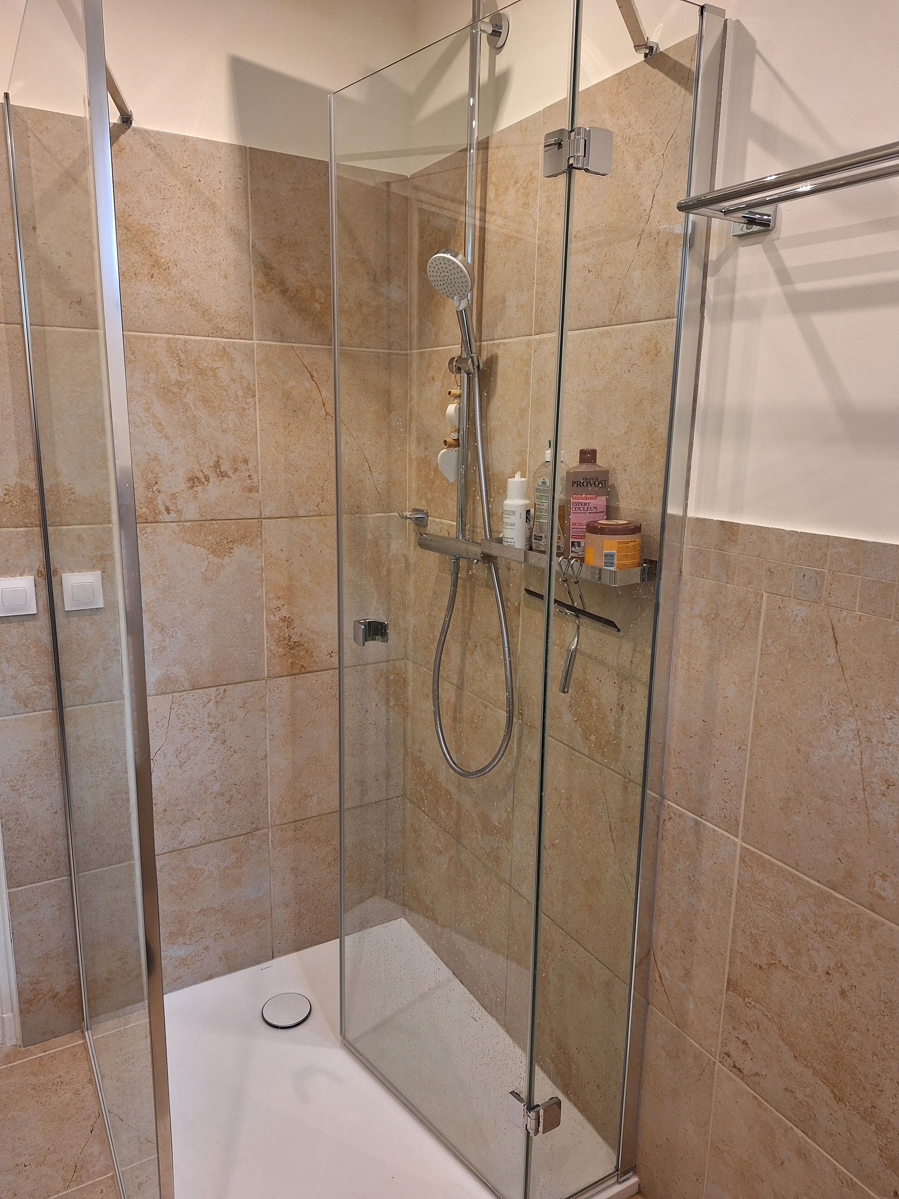 Cabina de ducha con paredes de vidrio, ducha de mano, grifo empotrado y repisa con artículos de aseo.
