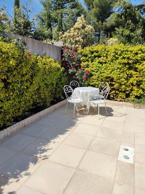Une terrasse extérieure avec une table et des chaises, entourée de haies et d’arbres bien entretenus.