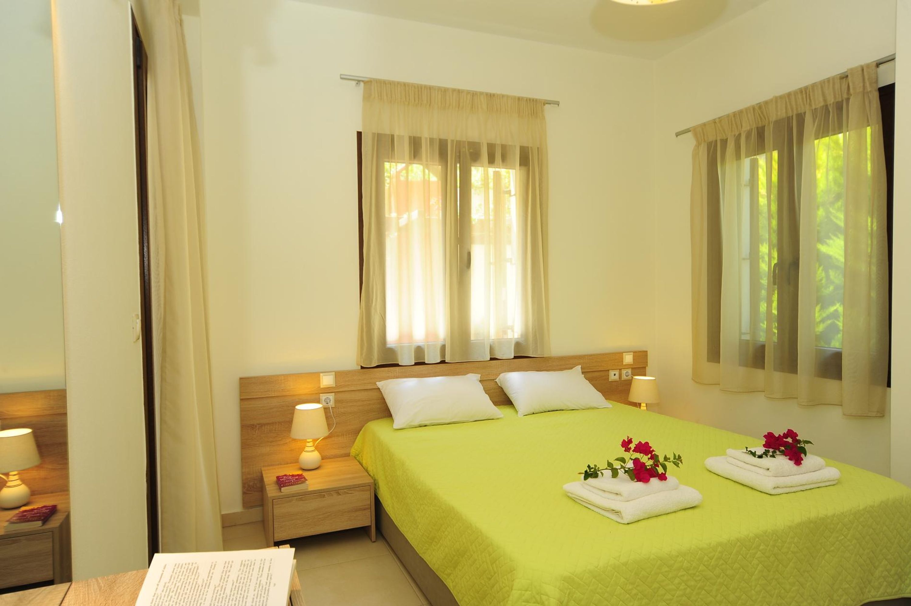 A hotel room with a large bed, two bedside tables with lamps, a window with curtains, and a view of greenery outside.
