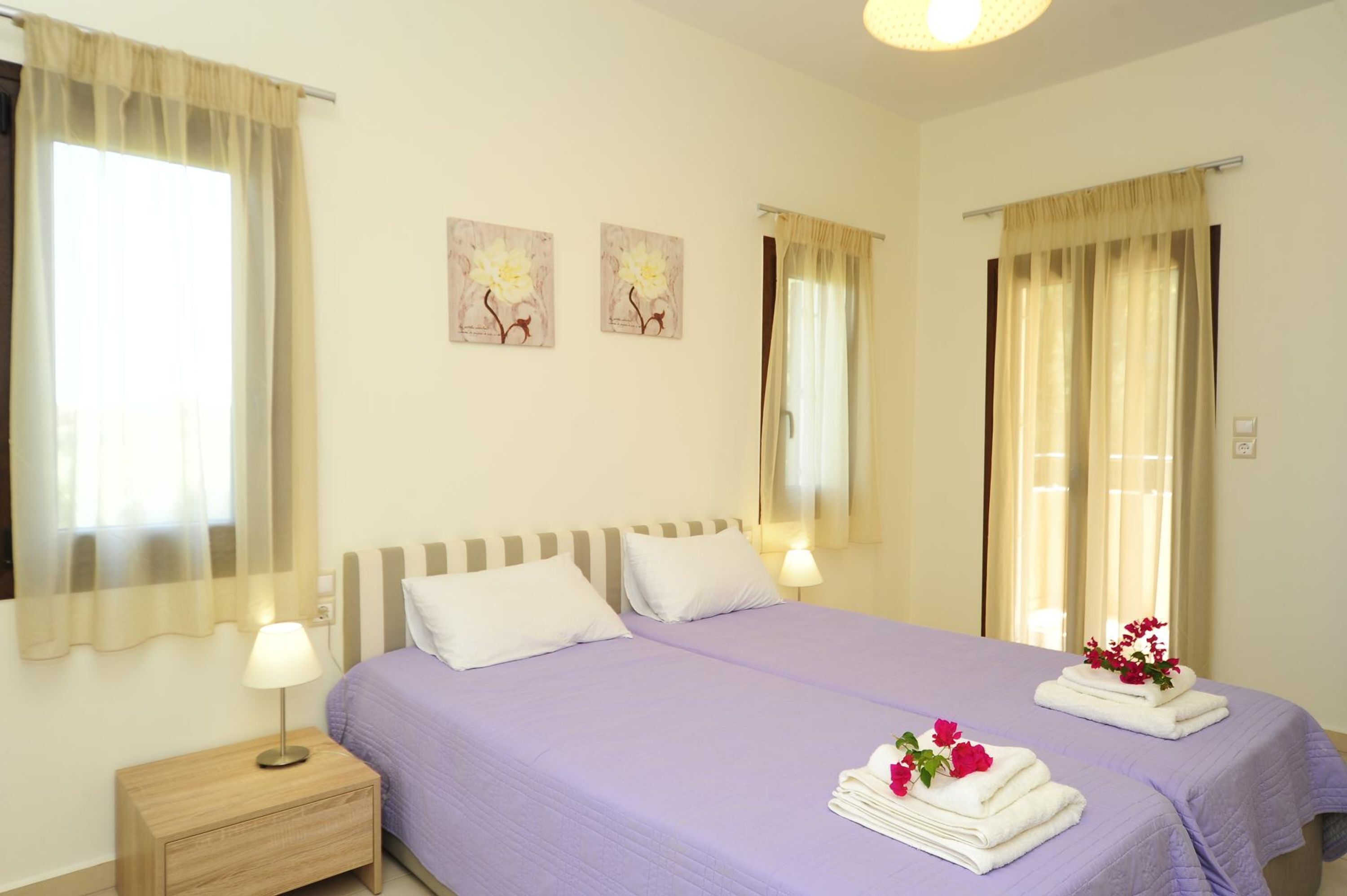 A hotel room with two beds, a nightstand, and a lamp. There are two paintings on the wall and a window with curtains.