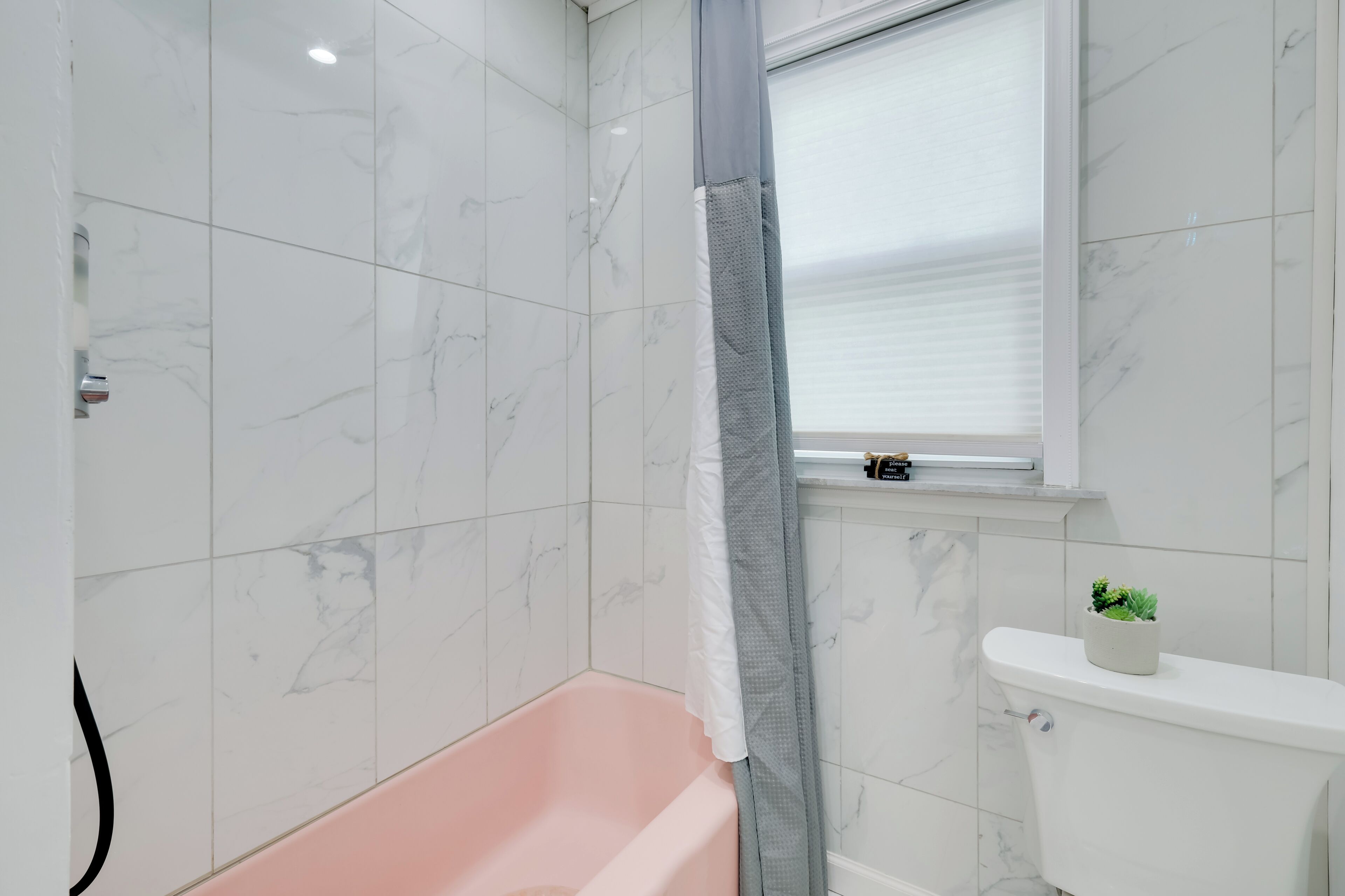 Et badeværelse med væg i marmor, et pink badekar, et toilet med en lille plante og et vindue med skodder.