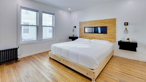 A bedroom with a wooden bed frame, white bedding, a mounted TV, and a small bedside table with a lamp.