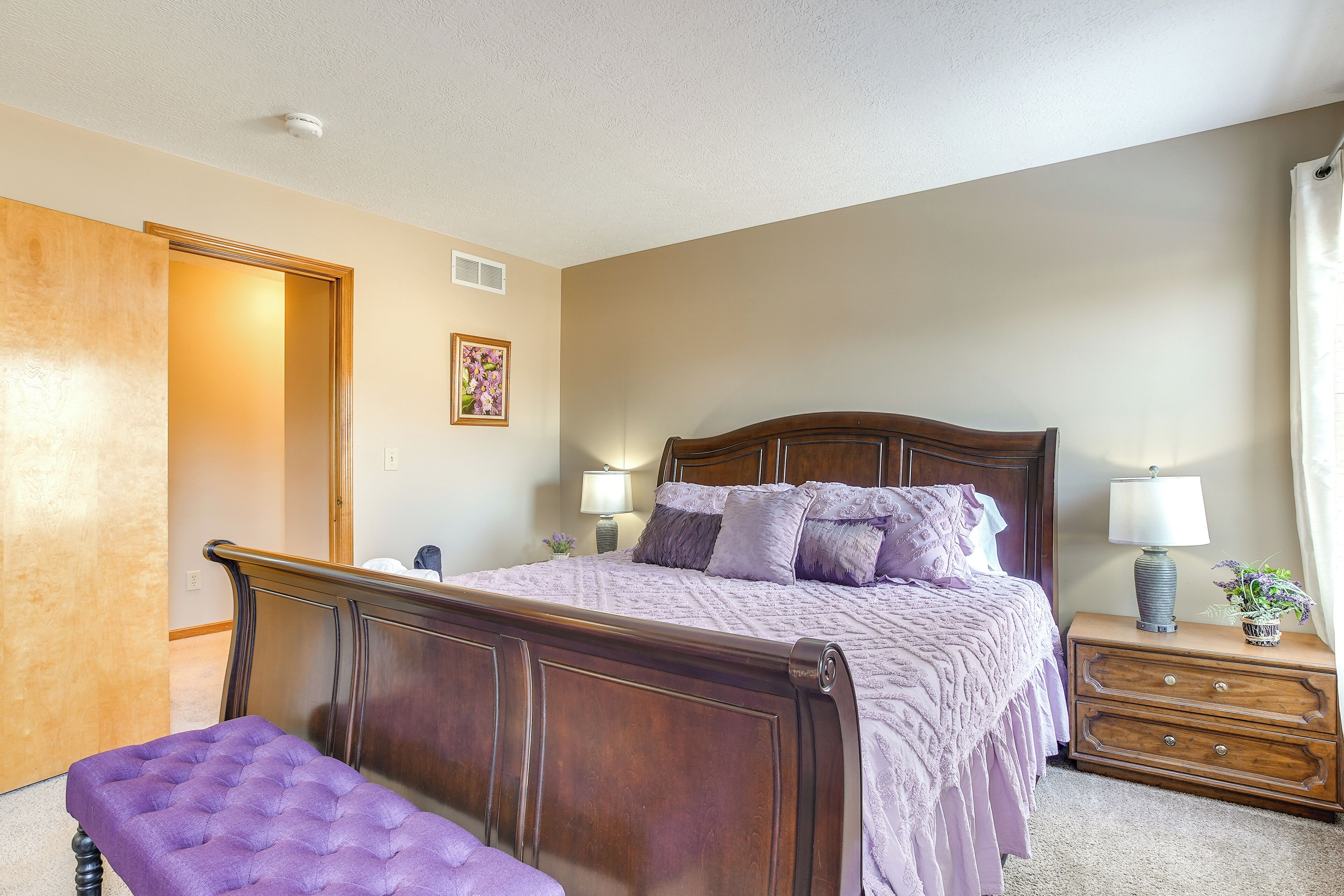 A bedroom with a wooden bed, a bench, a nightstand with a lamp, and a door leading to another room.