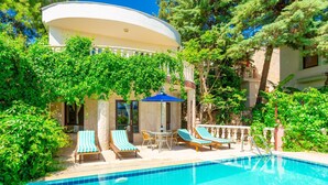 A pool area with lounge chairs, a table, and a building with a curved roof. - Kalkan Center 2+1 Villa in Nature - Variola (Antalya)