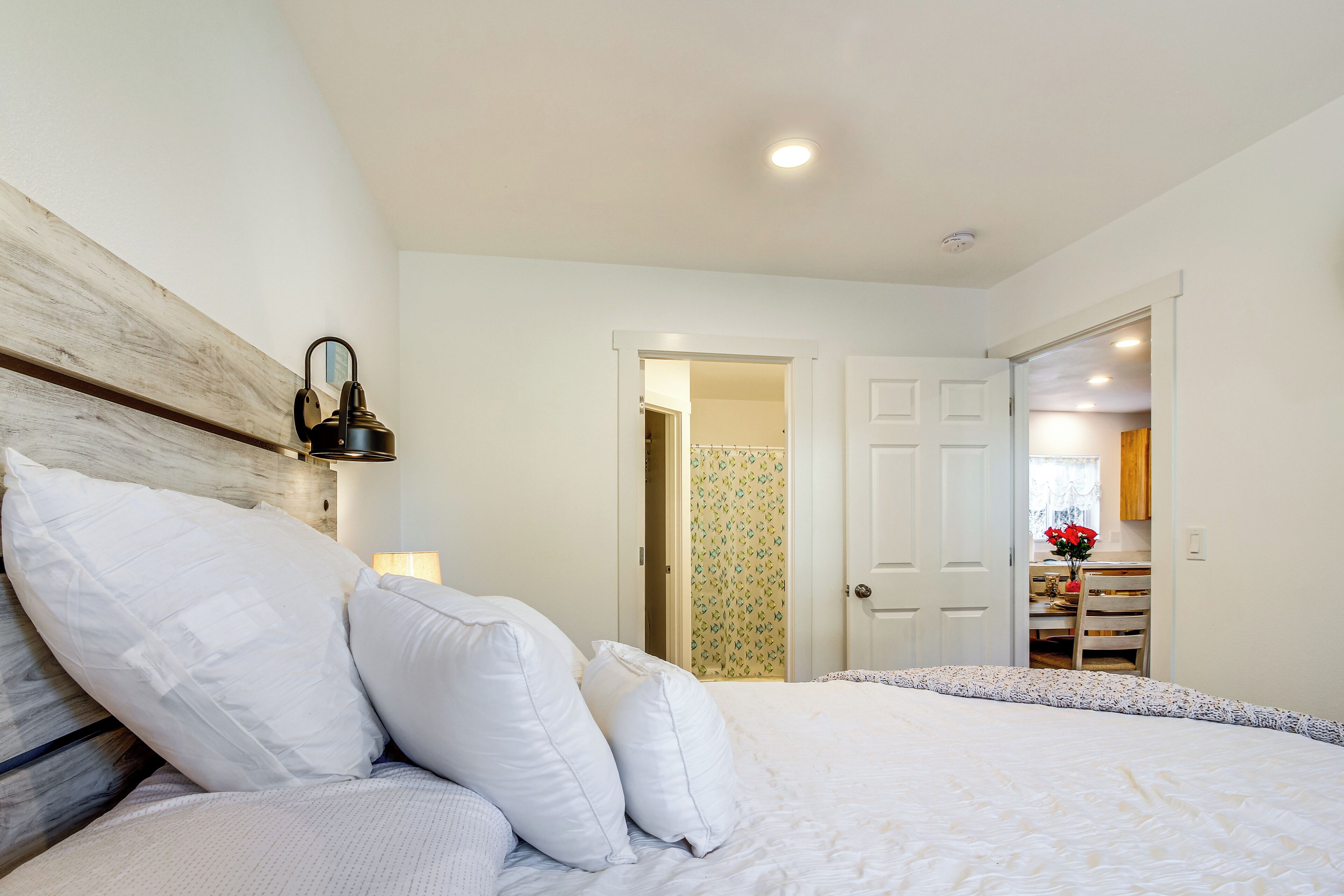 A bedroom with a large bed, white bedding, a wooden headboard, a nightstand with a lamp, and a door leading to a bathroom.