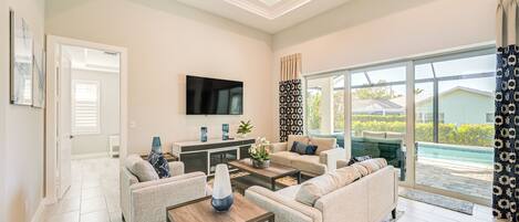 A modern living room with a flat-screen TV, comfortable seating, a coffee table, and a large window offering a view of a swimming pool.