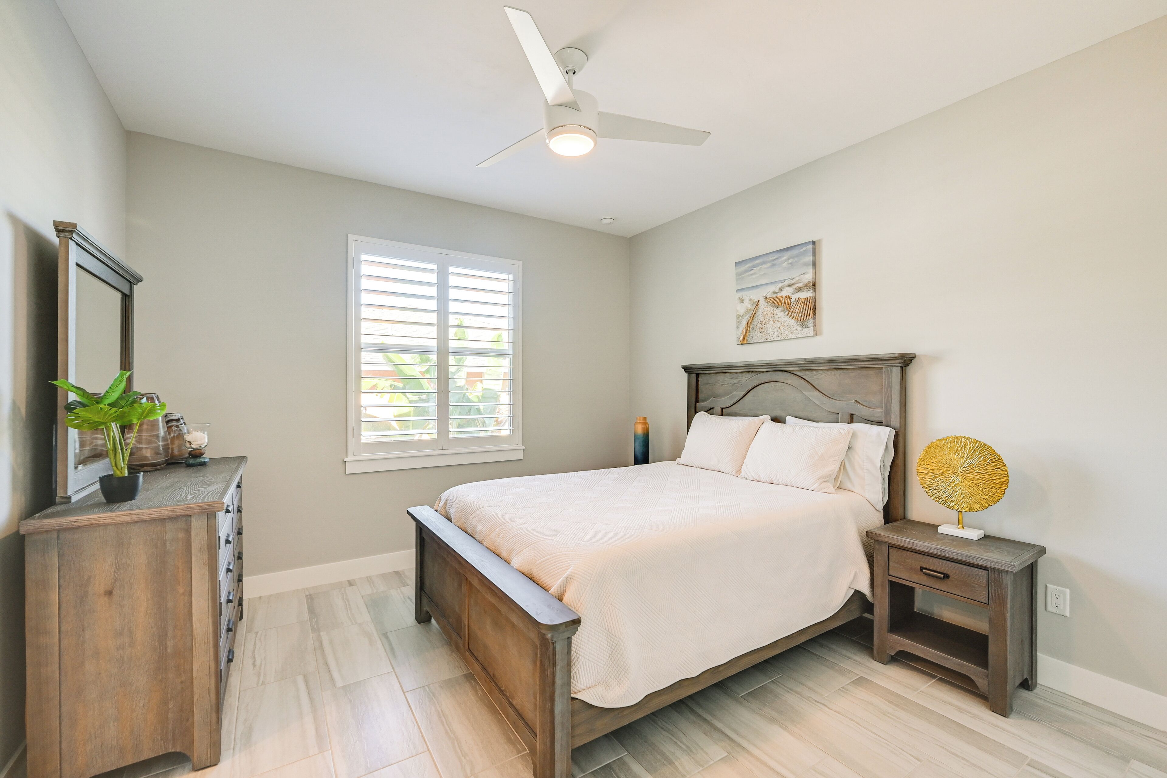 Une chambre à coucher comprenant un lit, une table de chevet, une commode, un ventilateur de plafond et une fenêtre avec des stores.