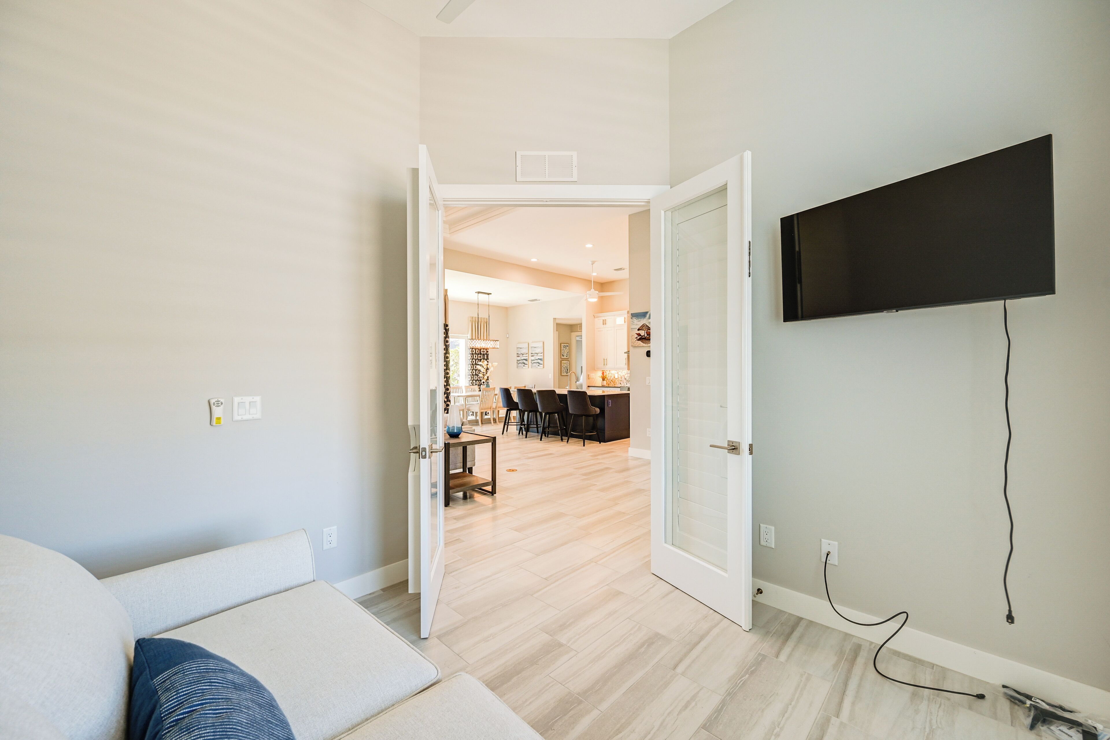 A modern living room with a sofa, a flat-screen TV mounted on the wall, and an open door leading to a dining area with chairs and a table.