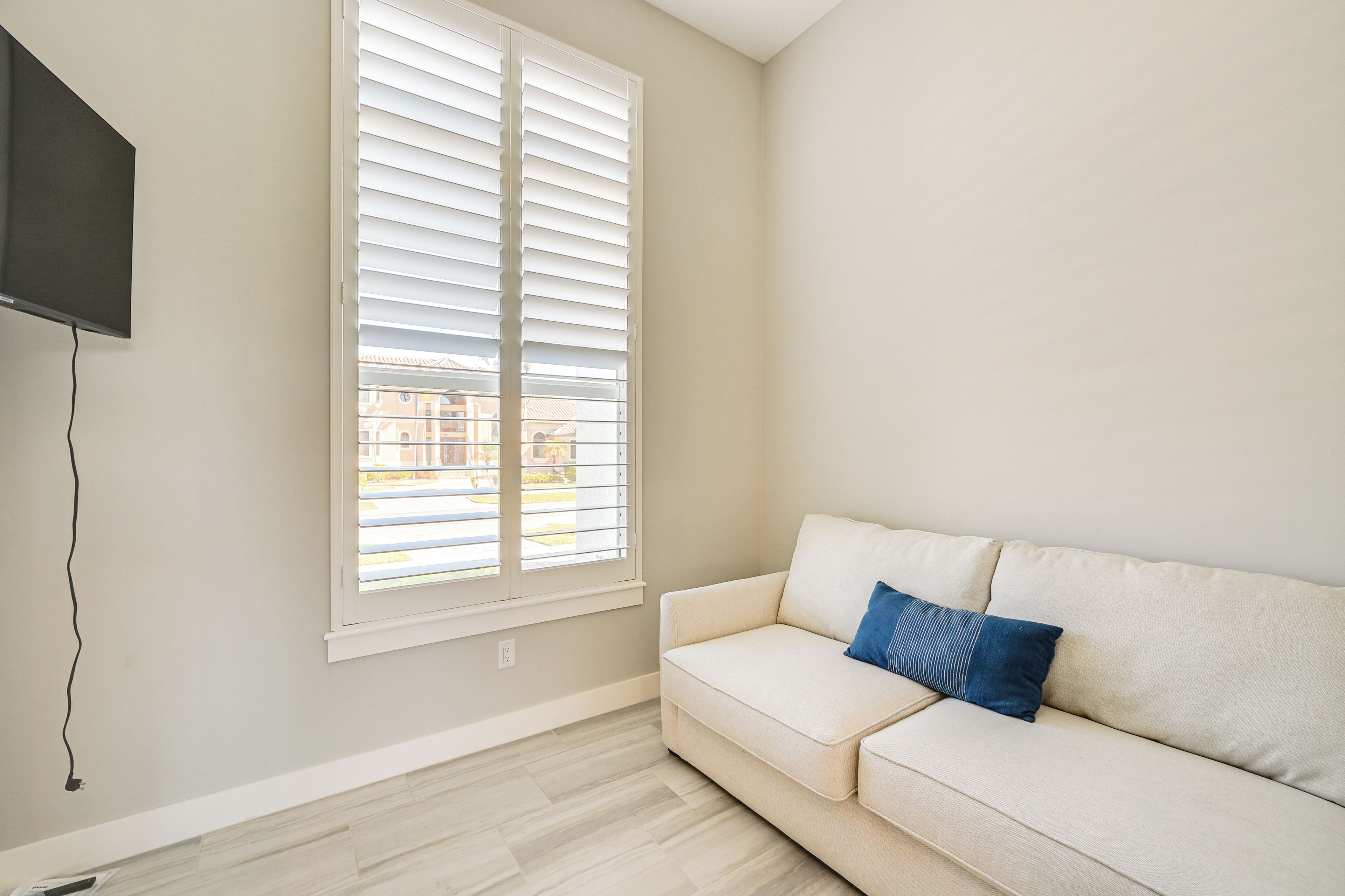 A living room with a beige sofa, a blue pillow, a TV mounted on the wall, and a window with blinds.