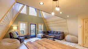 A cozy loft bedroom with a sloped ceiling, wooden beams, a large wooden table, a sectional sofa, and a view of the outdoors through skylights.