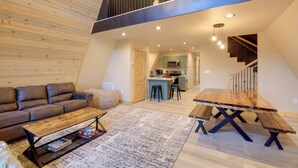 A modern loft-style living space with a wooden ceiling, a dining area with a wooden table and benches, a leather sofa, and a kitchen in the background.