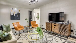 A modern living room with a sofa, two armchairs, a glass coffee table, a wooden TV stand, and a large flat-screen TV.