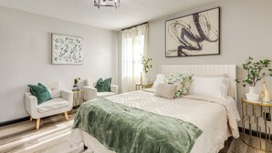A modern bedroom with a large bed, two armchairs, a small side table, and a decorative plant.