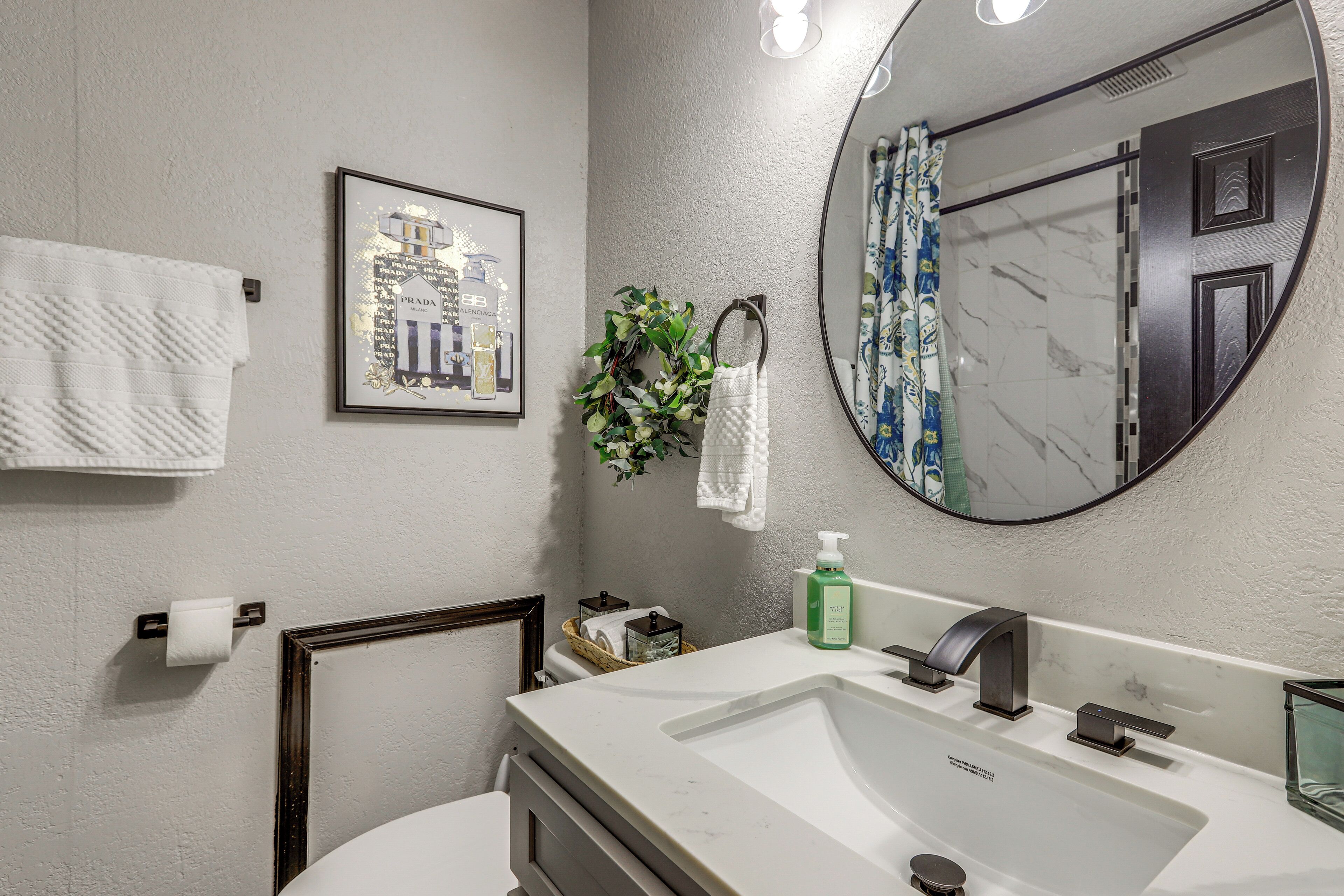 Ein Badezimmer mit weißem Waschbecken, einem Spiegel, einem gerahmten Bild, einem Handtuchhalter und einer Zimmerpflanze.