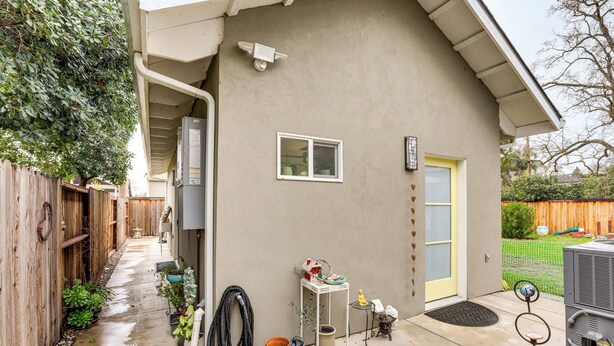 A residential backyard with a concrete patio, a wooden fence, potted plants, a small table with items, and a door with a yellow frame.